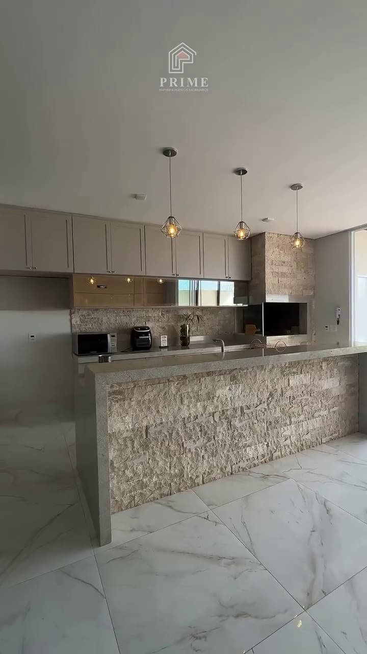 Área gourmet com ilha revestida em travertino e luminárias pendentes